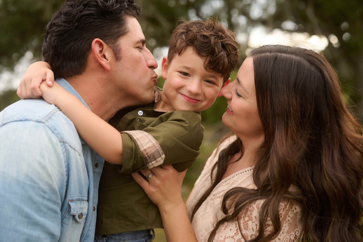 Parents kissing their smiling son beneath the Hill Country oaks
